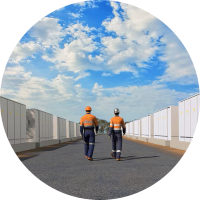 A GC image of two workers walking a path between a row of large batteries. The sky is blue and cloudy.